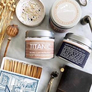 Könnte beinhalten: Eine Auswahl an Dekorationsartikeln, darunter Sojawachskerzen in Dosen mit den Aufschriften "Titanic" und "Scents of Titanic", eine Streichholzschachtel mit einer blau-weißen Illustration und ein kleines Notizbuch. Weitere Artikel sind eine kleine Schale mit goldenen Büroklammern, ein Stempel und Trockenblumen.