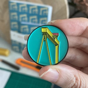 May include: A yellow and teal enamel pin featuring a stylized illustration of a yellow crane with the text "H&W" on the side.