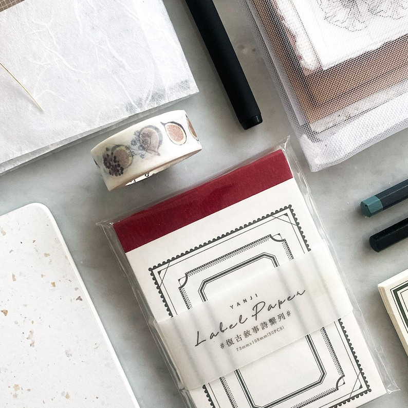 May include: A red and white notepad with a decorative border and the text "YANJI Label Paper # 73mmx108mm(32PCS)" on the front. The notepad is sitting on a white surface with other stationery items.