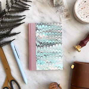 May include: A pink and blue marbled notebook with a pink spine. The notebook is on a white marble surface with a fern, a pen, scissors, a dried poppy pod, and a brown leather journal.