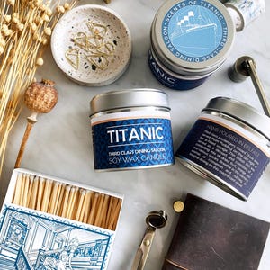 Könnte beinhalten: Draufsicht auf Artikel zum Thema Titanic. Silberne Dosen mit Sojawachskerzen, eine Streichholzschachtel mit einer blauen Illustration, eine kleine Schale mit goldenen Büroklammern und ein ledergebundenes Notizbuch mit einem Schiffsrad-Anhänger.