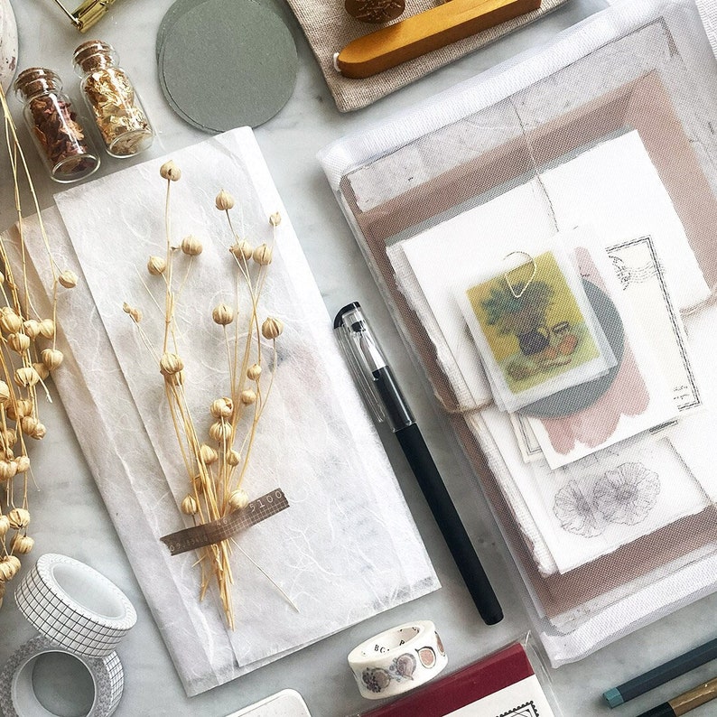 May include: A white paper envelope with a bundle of dried beige flowers and a brown label with the text "3100". The envelope is on a white surface with other craft supplies, including a pen, a roll of washi tape, and a small glass bottle with gold flakes.