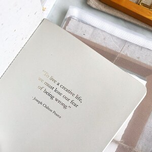 May include: A close-up of a page in a notebook with a quote in gold lettering: "To live a creative life, we must lose our fear of being wrong." - Joseph Chilton Pearce