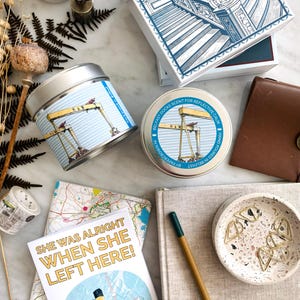 Może przedstawiać: Asortyment prezentów i artykułów papierniczych. Obejmuje ozdobne pudełko z niebiesko-białą ilustracją, świecę w puszce z motywem żurawia oraz kartkę z napisem "SHE WAS ALRIGHT WHEN SHE LEFT HERE!". Inne przedmioty to notatnik, mała miseczka ze spinaczami i skórzany portfel.