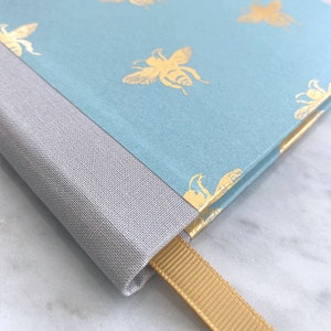 Op de afbeelding: Een lichtblauw notitieboek met een gouden bijenpatroon en een grijze stoffen rug. Het notitieboek heeft een gouden lint als bladwijzer.