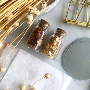 May include: Two small glass bottles with cork tops. One bottle contains dried rose petals and the other contains gold leaf. The bottles are on a white surface with a stack of gray paper circles.