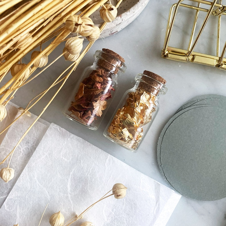 May include: Two small glass bottles with cork tops. One bottle contains dried rose petals and the other contains gold leaf. The bottles are on a white surface with a stack of gray paper circles.
