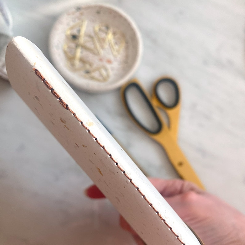 May include: Close-up of the edge of a white, speckled, rectangular tray with a copper-colored stitched border.