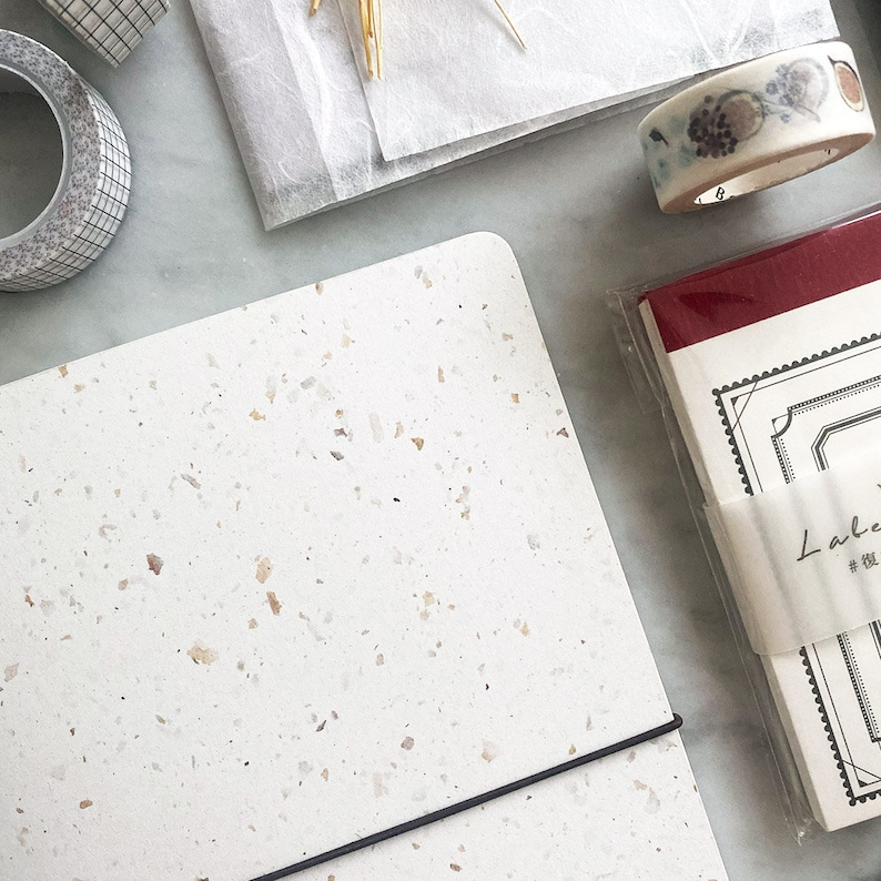 May include: A white notebook with a speckled texture and a black elastic band closure. The notebook is lying on a white surface with other stationery items.