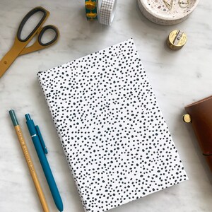 May include: A white notebook with a black polka dot pattern. The notebook has a black elastic closure and a black ribbon bookmark. The notebook is lying on a white marble surface.
