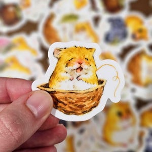 May include: A sticker featuring a smiling hamster in a woven basket. The hamster has orange and white fur, with a happy expression. The basket is brown, and the sticker has a white border. The background shows other similar stickers.