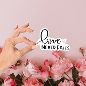 May include: A white sticker with black lettering that reads "love never fails" with a small pink heart. The sticker is held by a hand against a pink background with pink roses.