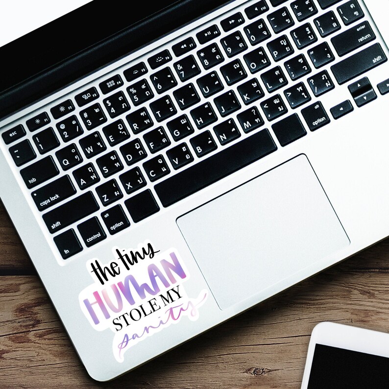 May include: A laptop computer with a sticker on the trackpad that says "the tiny HUMAN STOLE MY sanity". The sticker is a gradient of pink and purple.