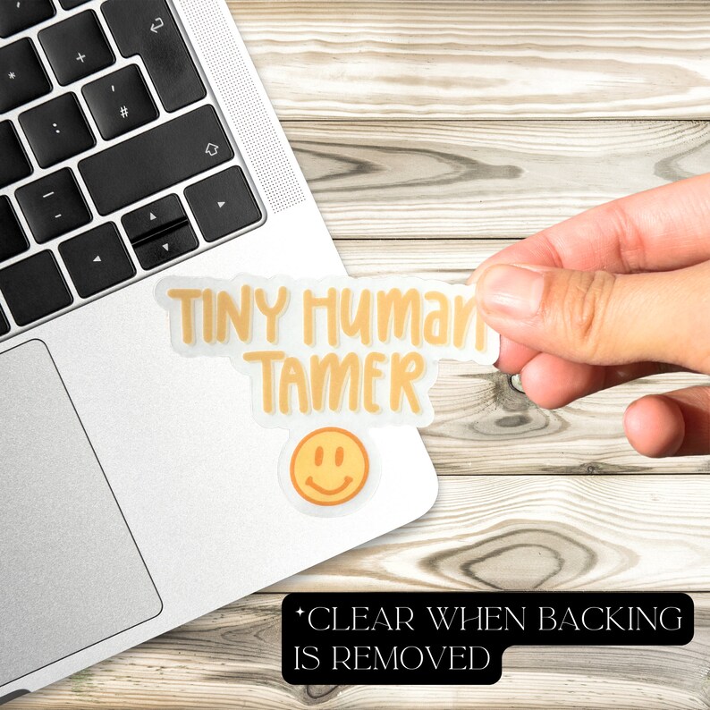May include: A yellow sticker with the text "TINY HUMAN TAMER" and a smiley face. The sticker is on a laptop computer. The text "CLEAR WHEN BACKING IS REMOVED" is printed below the sticker.