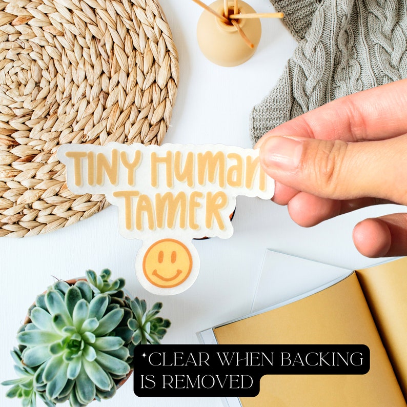 May include: A yellow sticker with the text "Tiny Human Tamer" and a smiley face. The sticker is being held by a hand. The text "CLEAR WHEN BACKING IS REMOVED" is visible below the sticker.