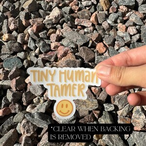 May include: A white sticker with the text "Tiny Human Tamer" in yellow lettering. A yellow smiley face is below the text. The sticker is on a background of small gray and pink rocks.