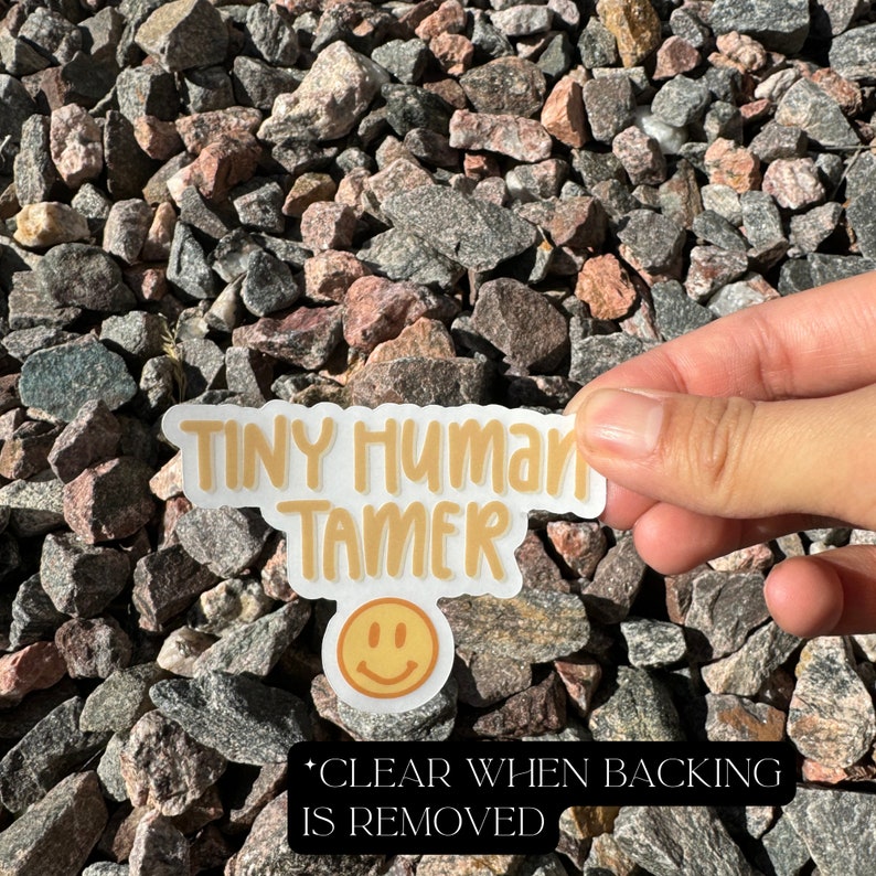 May include: A white sticker with the text "Tiny Human Tamer" in yellow lettering. A yellow smiley face is below the text. The sticker is on a background of small gray and pink rocks.