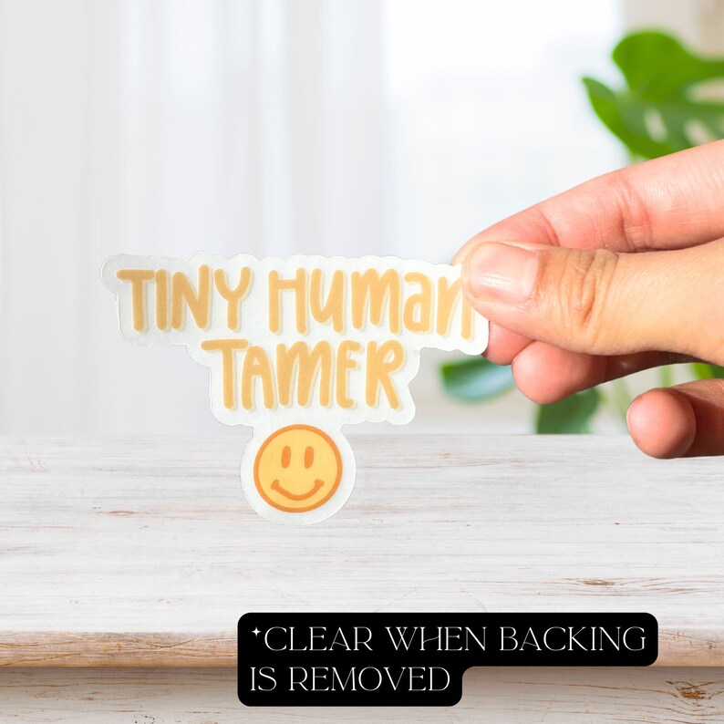 May include: A yellow sticker with the text "Tiny Human Tamer" and a smiley face. The sticker is being held by a hand.