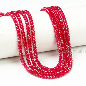 May include: A collection of faceted, bright red beads strung together, displayed on a white cylindrical surface. The beads are arranged in multiple strands, showcasing their color and texture. The image is well-lit, highlighting the beads' reflective qualities.
