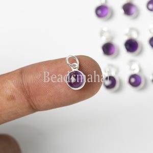 Può includere: Un piccolo ciondolo rotondo di pietra preziosa viola con una cauzione color argento. Il ciondolo è tenuto da un dito, con altri ciondoli simili visibili sullo sfondo. Il testo "Beadsmaha" è parzialmente visibile.