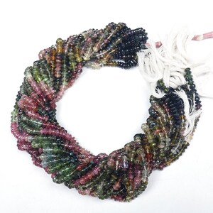 May include: A strand of multi-colored tourmaline beads. The beads are faceted and have a variety of colors, including green, pink, black, and brown. The beads are strung on a white cord.