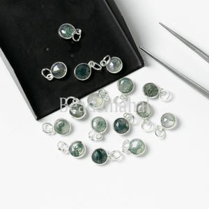 Puede incluir: Una colección de 15 dijes de plata con piedras de ágata verde musgo. Los dijes están esparcidos sobre una superficie blanca.