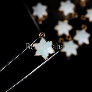May include: A close-up of a white star-shaped pendant with a gold-coloured border. The pendant is held by tweezers against a black background. Several similar pendants are visible. The text "Beadsaha" is partially visible.