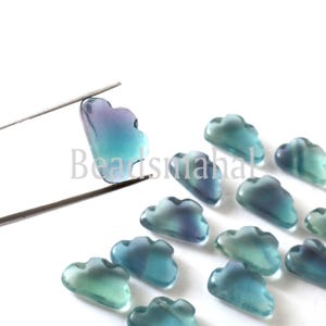 May include: A collection of blue and green glass cloud shaped beads. The beads are translucent and have a gradient of color from blue to green. The beads are arranged in a row on a white surface.