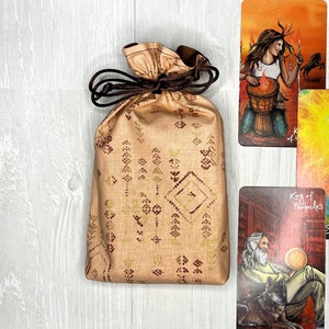 May include: A tan fabric tarot card bag with a brown drawstring, next to three tarot cards. The bag has a geometric pattern in brown and gold. The tarot cards feature colorful illustrations of figures and symbols.