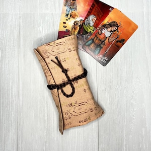 Puede incluir: Una bolsa de tela marrón con un patrón geométrico y un cierre de cordón marrón. La bolsa está enrollada y se encuentra sobre una superficie de madera blanca. Se pueden ver tres cartas de tarot en el fondo, con ilustraciones de personas y animales.