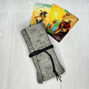 Könnte beinhalten: Eine graue Tarotkarten-Tasche aus Stoff mit einem schwarzen Band und einer Quastenverschlusses. Die Tasche ist mit einem sich wiederholenden Muster aus kleinen weißen Punkten verziert. Im Hintergrund sind drei Tarotkarten zu sehen, die farbenfrohe Illustrationen und den Text "The Emperor" zeigen.