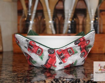 May include: A white ceramic bowl with a fabric cozy featuring a pattern of red trucks, green trees, and snowflakes. The cozy has a green trim. The bowl is on a speckled countertop.