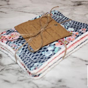 以下が含まれることがあります： ひもと茶色のタグで結ばれた、模様入りの布の正方形のスタック。生地は、ネイビーブルーのオープン織りデザイン、ピンクとブルーの花柄、白い縁が特徴です。タグは空白です。