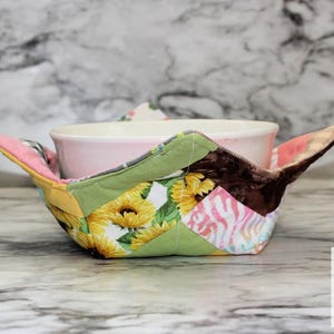Puede incluir: Un cuenco de cerámica rosa se encuentra dentro de un colorido y acolchado protector de cuenco. El protector presenta un diseño de retazos con girasoles, tela verde y detalles en rosa y marrón. El protector tiene solapas extendidas para facilitar su manejo.