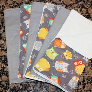 May include: A stack of baby burp cloths in shades of grey and patterned fabric. The cloths feature a grey background with colourful owl designs in orange, yellow, green, and white. One cloth is white and textured. The cloths are neatly stacked.