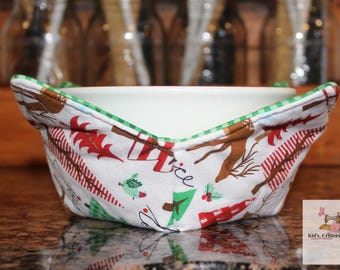 May include: A white bowl cozy with a Christmas-themed pattern featuring red, green, and brown festive designs. The cozy has a green gingham trim and is designed to hold a bowl. The background is blurred, showing bottles and a wooden surface.