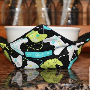 May include: A black bowl cozy with a pattern of white, light blue, and lime green video game controllers. The cozy is folded to hold a bowl and has a square shape with extended corners.