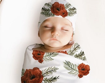 May include: A baby wrapped in a white blanket and matching hat, featuring a red poppy and green leaf design. The baby lies in a woven basket. The blanket and hat are decorated with a floral pattern.