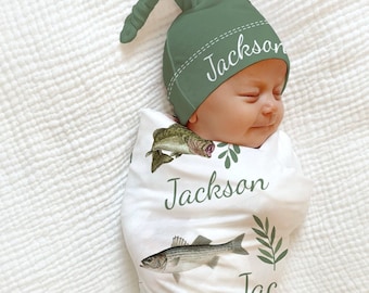 May include: A baby swaddled in a white blanket with fish illustrations and the name "Jackson" printed on it. The baby wears a green hat with the name "Jackson" on it. The words "Swaddle + Hat" are at the bottom.