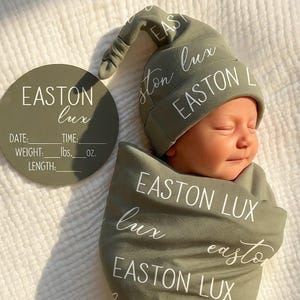 May include: A newborn baby wrapped in a sage green swaddle blanket and matching hat, both printed with the name "EASTON LUX" in white script. A circular sign with the same name and fill-in-the-blank details is also present.