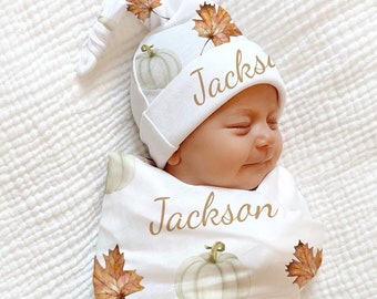 May include: A white swaddle blanket with a fall theme featuring pumpkins and leaves. The blanket has the name "Jackson" written on it multiple times. A white hat with a pointed tip and the name "Jackson" written on it is on the baby's head.
