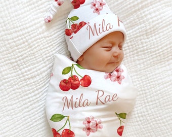 May include: A white swaddle blanket with a cherry and floral print. The blanket has the name "Mila Rae" written on it. A white hat with a cherry and floral print and the name "Mila" written on it is on top of the swaddle.