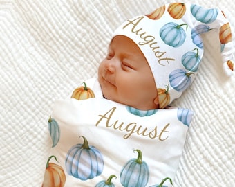 May include: A baby swaddled in a white blanket and wearing a matching hat, both adorned with watercolor pumpkins in shades of blue, orange, and green. The word "August" is written in gold script on both items.