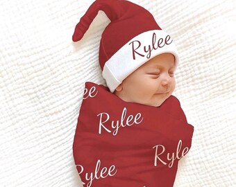 May include: A red swaddle blanket with the name "Rylee" printed on it. A baby is wearing the swaddle and a matching red Santa hat with a white brim.