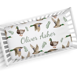 May include: White crib sheet with a green and brown duck pattern. The sheet has the name "Oliver Asher" printed in green.