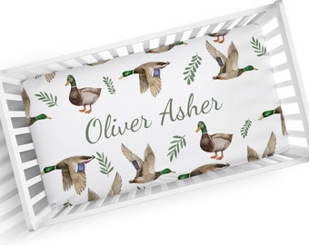 May include: White crib sheet with a green and brown duck pattern. The sheet has the name "Oliver Asher" printed in green.