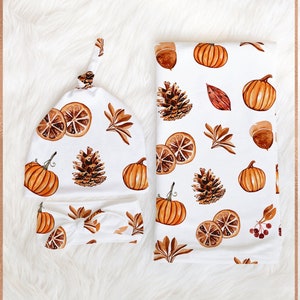 May include: A white knotted hat and headband with a fall-themed print featuring pumpkins, pinecones, leaves, and orange slices. The text "gray THE LABEL" is printed on the top of the image.