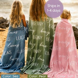 May include: Three personalized blankets in sea, pear, and soft colors are displayed on a sandy beach. Each blanket is draped over a person and features a name in a white script font. The sea blanket has the name "Lucas", the pear blanket has the name "Benny", and the soft blanket has the name "Avery".