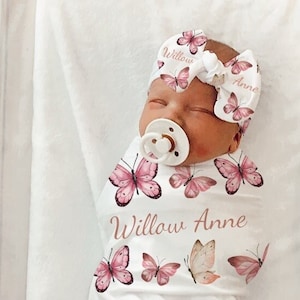 Peut inclure: Une couverture blanche pour bébé avec des papillons roses et le nom "Willow Anne" répété partout. Un bébé est emmailloté dans la couverture et porte un bandeau rose avec le même nom.