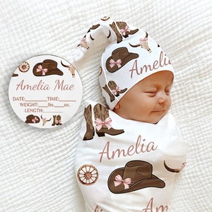 May include: A white swaddle blanket with a pink and brown cowboy boot, hat, and skull pattern. The blanket has the name "Amelia" written on it in pink. A matching pink and brown cowboy hat with the name "Amelia" written on it is on a baby's head.